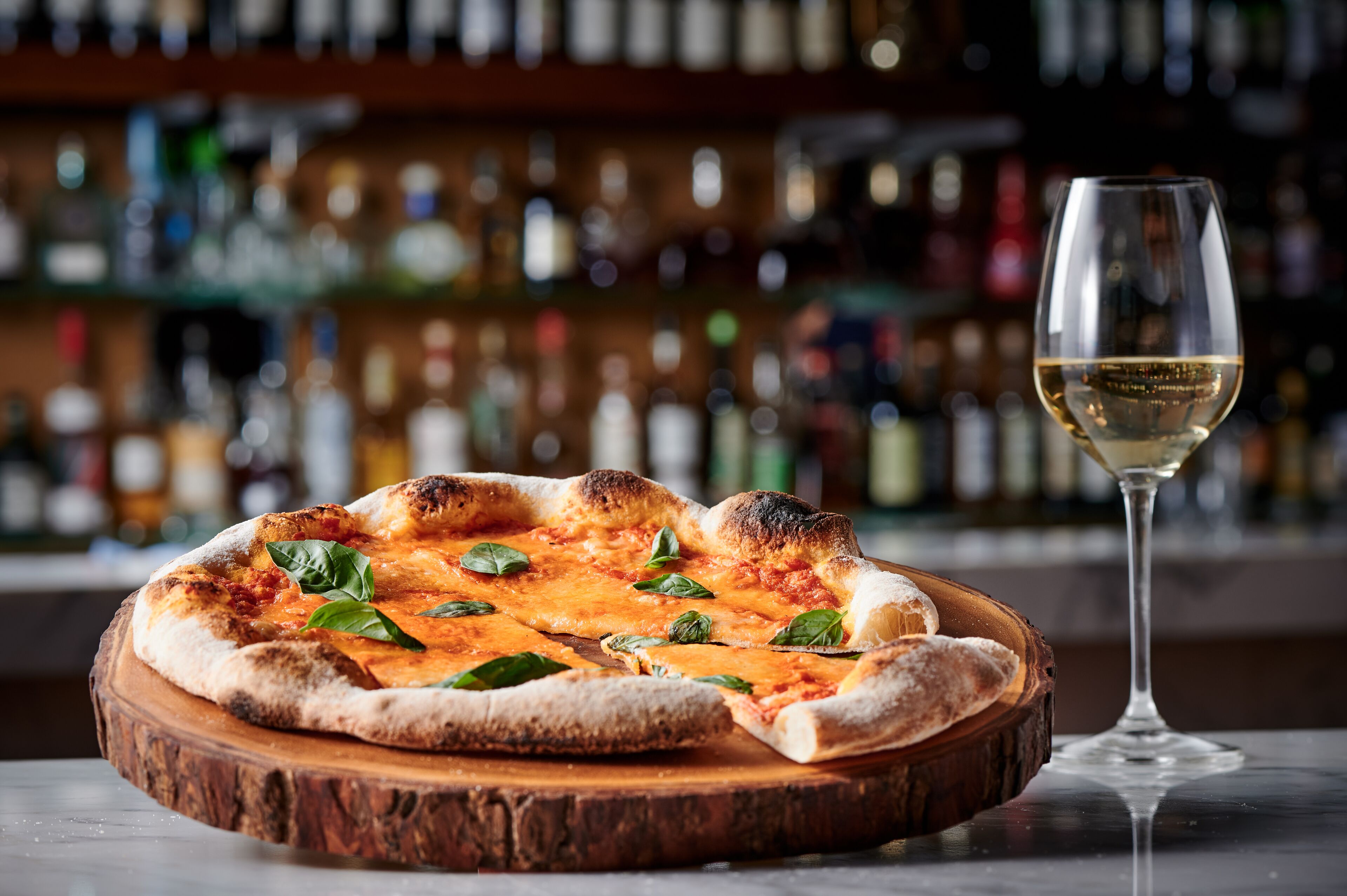 A pizza on a wooden board with a glass of white wine beside it.