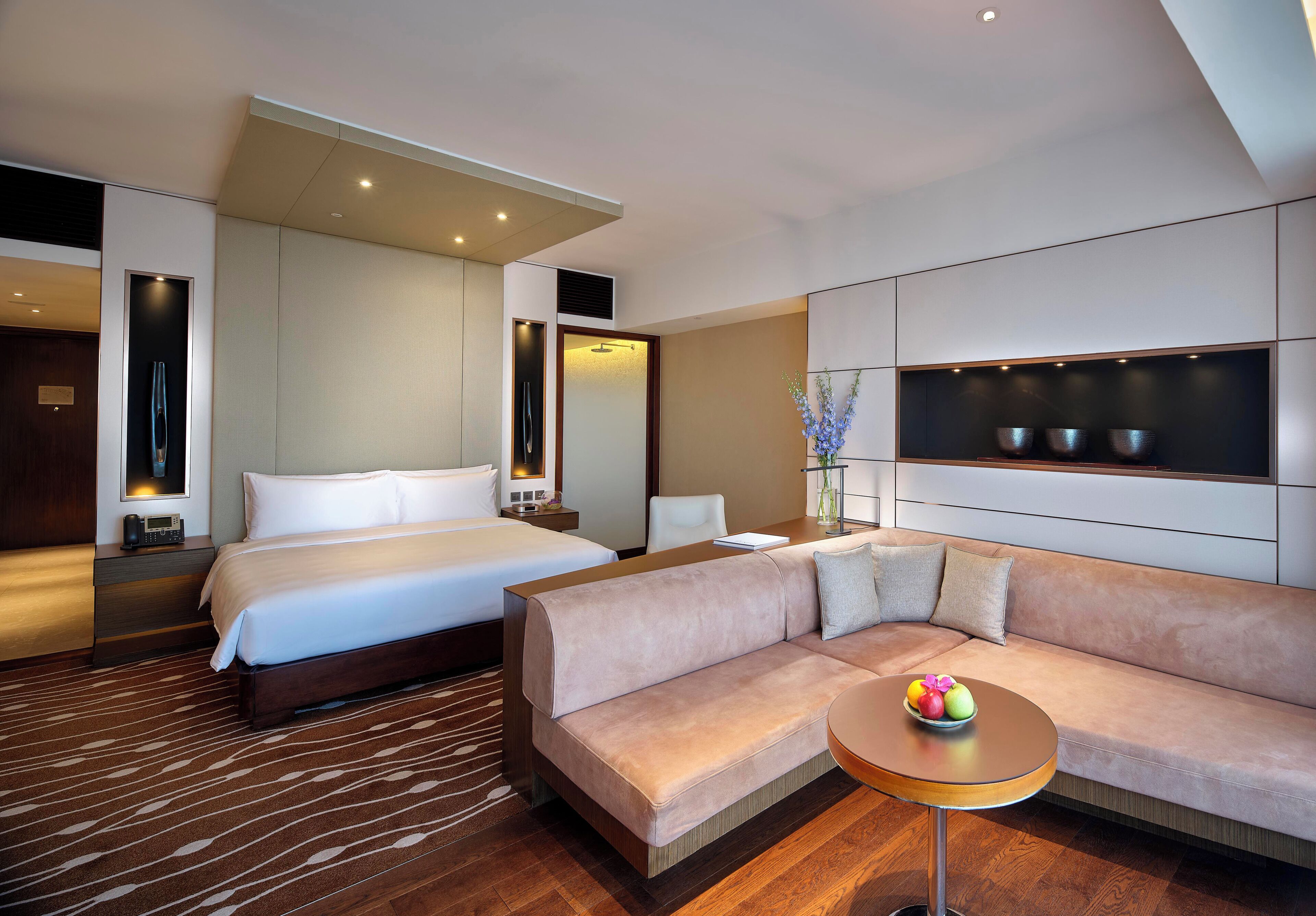 A modern hotel room with a bed, a sofa, a small table with fruit, and a TV.