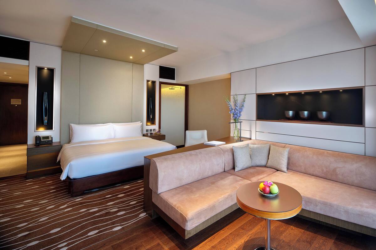 Une chambre d’hôtel moderne avec un grand lit, un canapé, une petite table avec des fruits et un téléviseur fixé au mur.