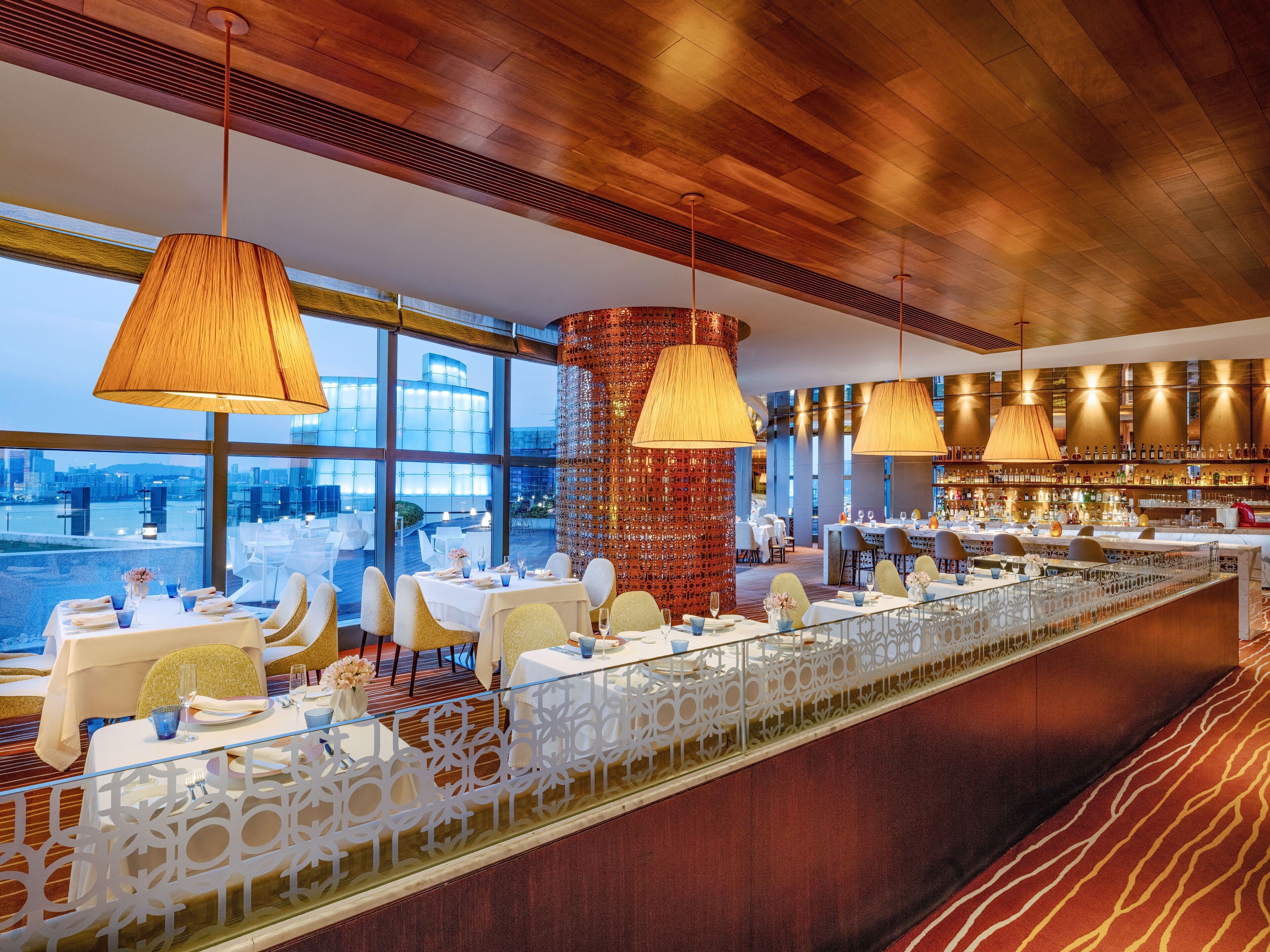 Un restaurante moderno con una larga mesa de comedor, múltiples lámparas colgantes y vistas de la ciudad a través de amplios ventanales.