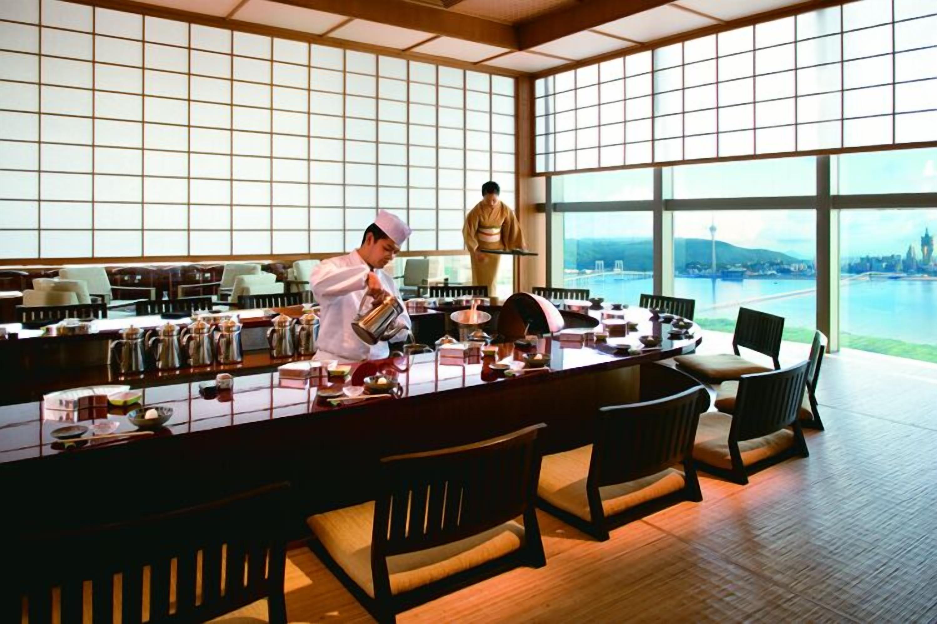 Un restaurant japonais traditionnel avec une grande table en bois, des tatamis et une vue sur le paysage urbain grâce à de grandes fenêtres.
