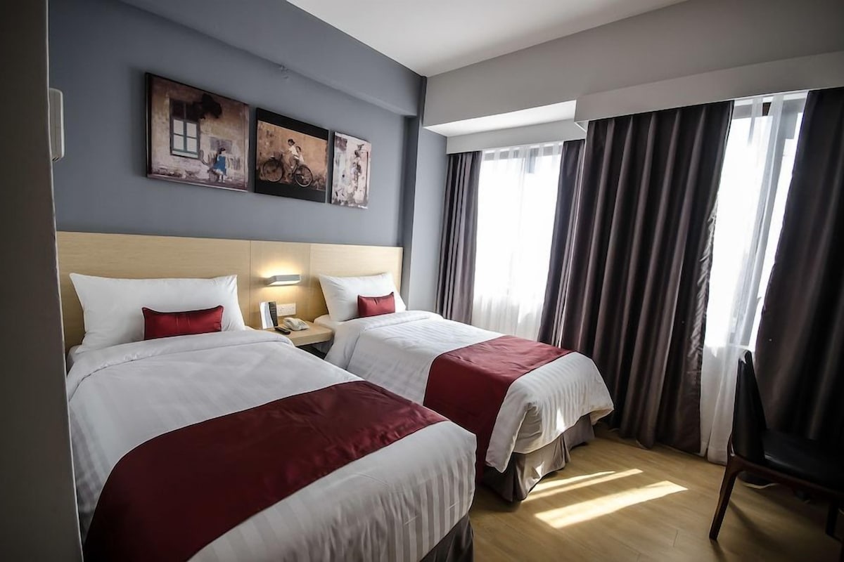A hotel room with two beds, a wooden headboard, a nightstand, a chair, and a window with curtains.