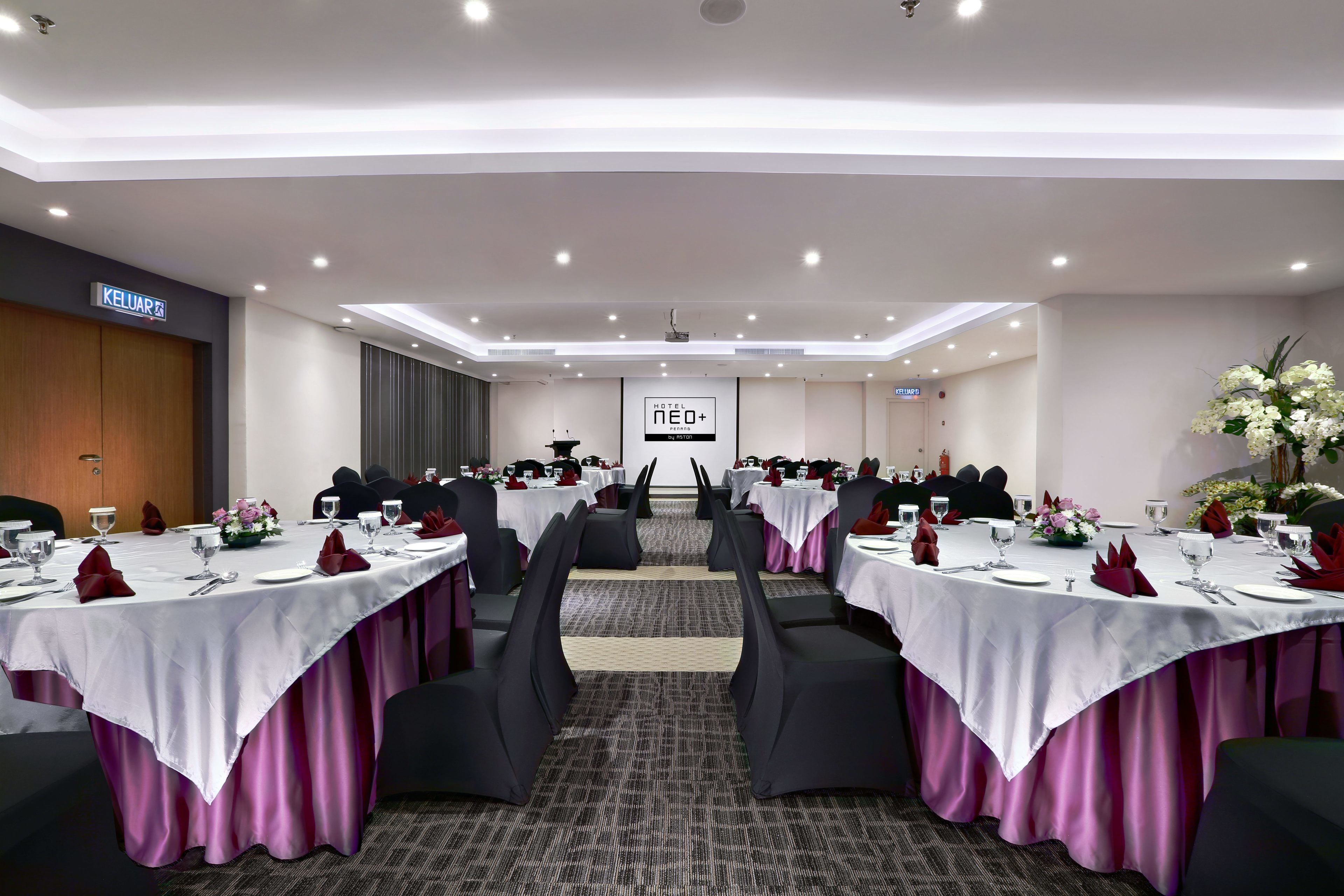Une salle de conférence d’hôtel avec plusieurs tables dressées pour un repas, un pupitre et une enseigne indiquant « HÔTEL NEO+ ».