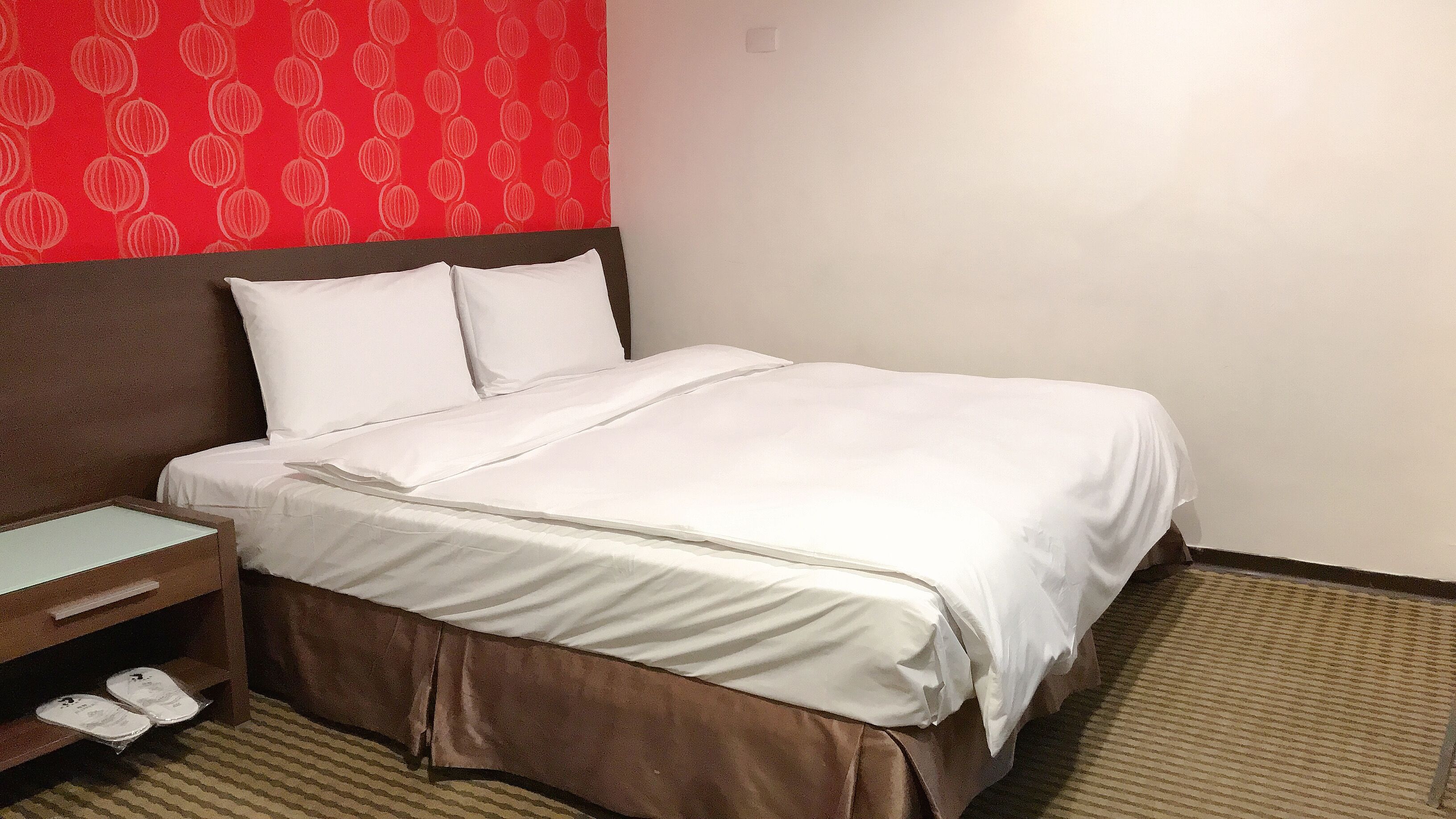 A hotel room with a neatly made bed, a wooden nightstand with a lamp, and a red patterned wall.