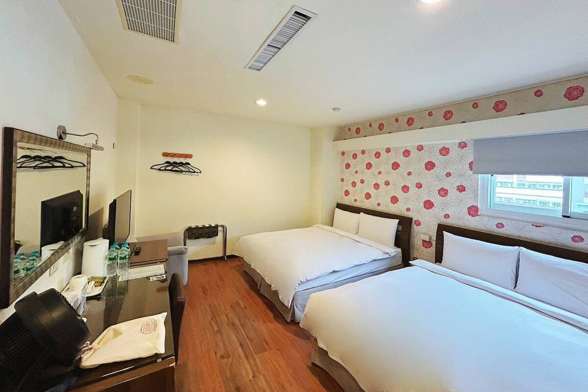 A hotel room with two beds, a desk with a chair, a mirror, a small table with toiletries, and a window with a view.