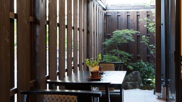 Una terraza de madera con una mesa, sillas, una planta en maceta y vistas a un jardín con un árbol y rocas.