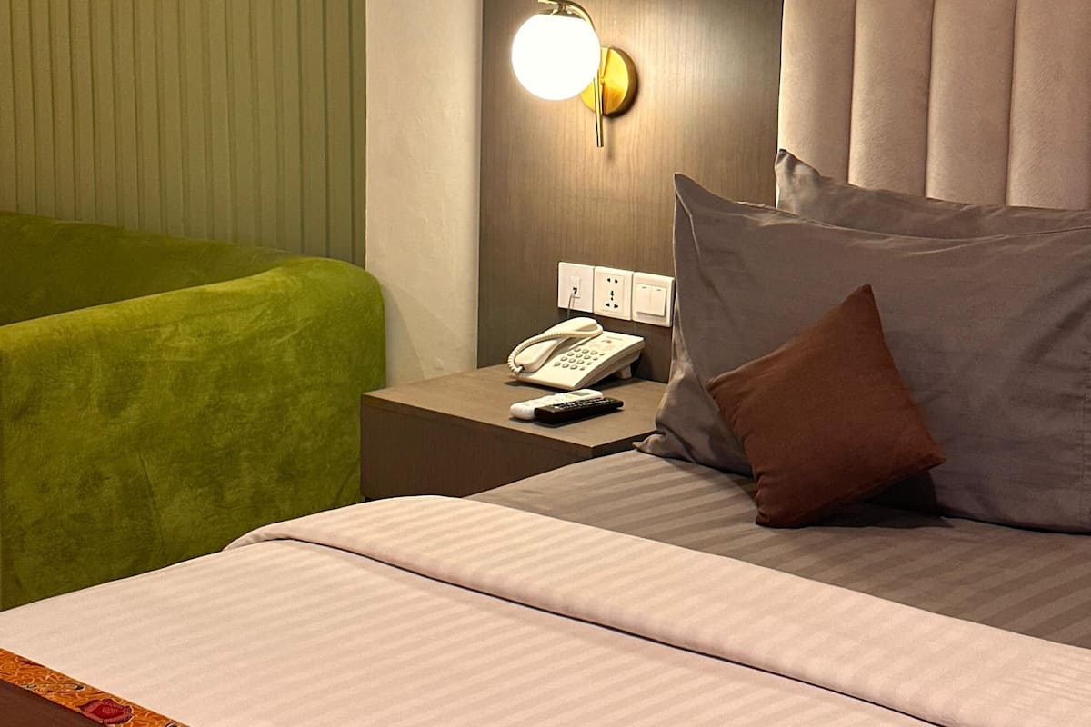 Ein Hotelzimmer mit einem Bett, einem Nachttisch mit Telefon und Fernbedienung, einem grünen Sessel und einer Wandleuchte.