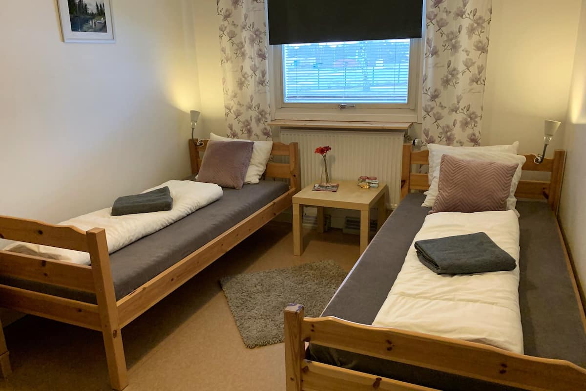 A room with two wooden beds, a small table with a vase and books, and a window with blinds.