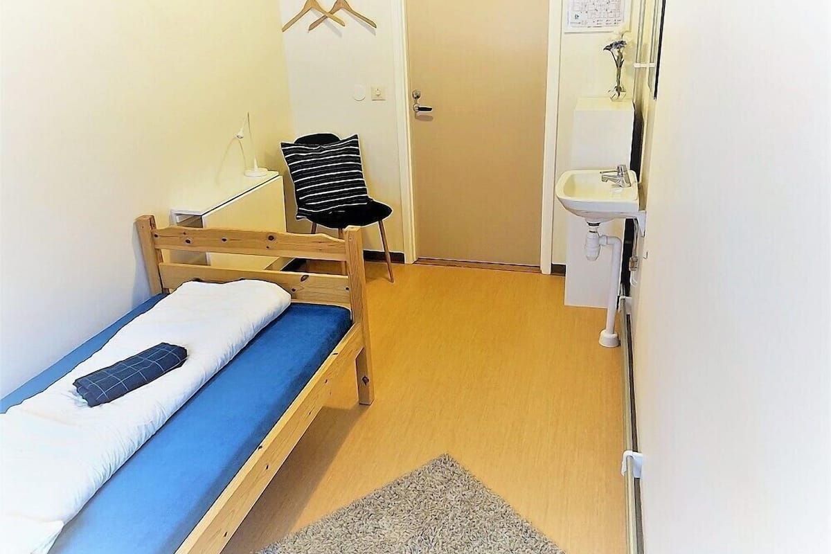 A small, single-bed room with a wooden bed frame, a blue mattress, a black and white striped chair, a white sink, and a door leading to another room.
