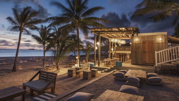 Un resort frente a la playa con mobiliario de madera en terraza, palmeras y un espacio al aire libre cubierto con mesas y sillas.