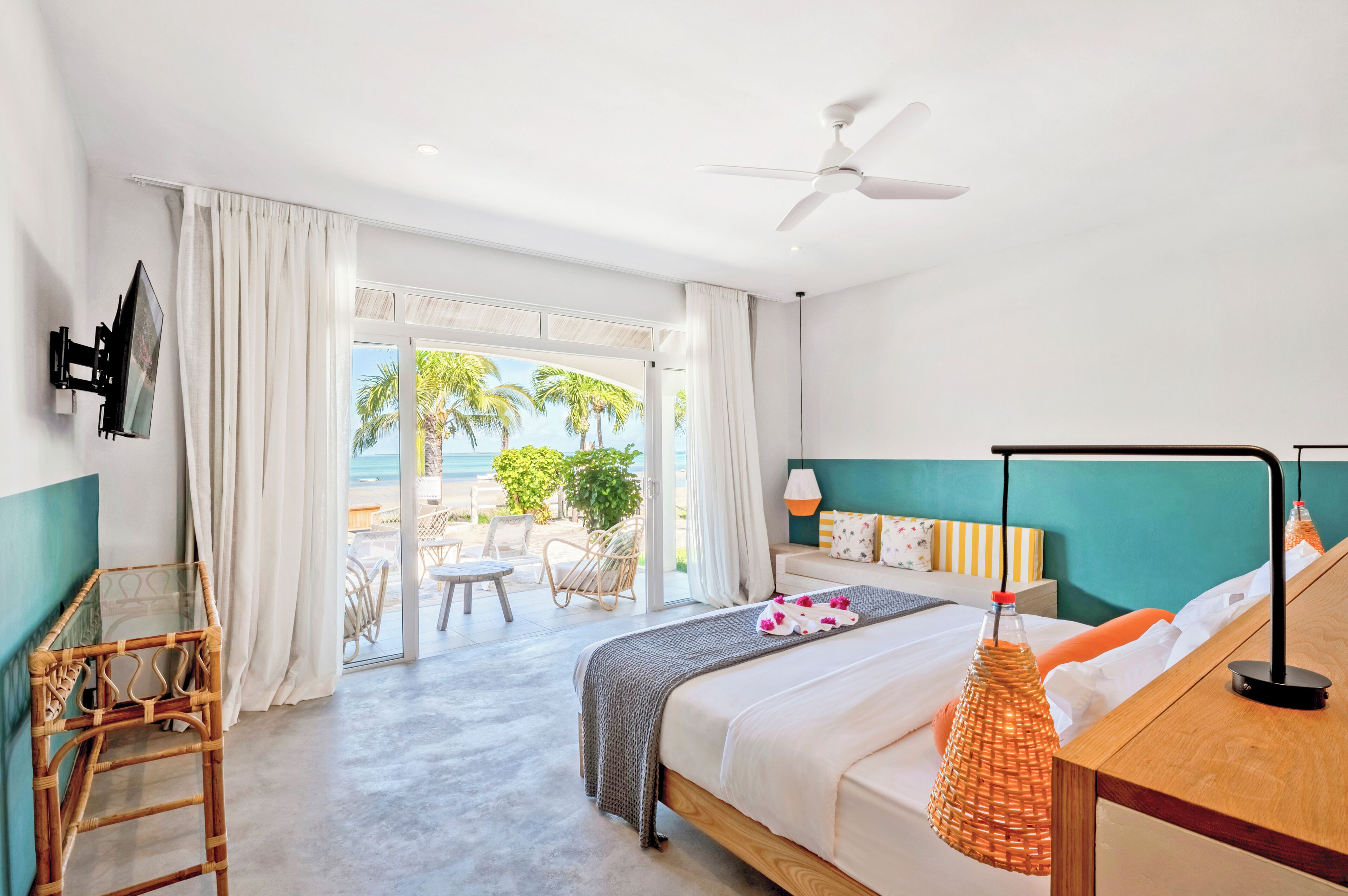 A hotel room with a bed, a desk, a chair, a TV, and a view of the beach.