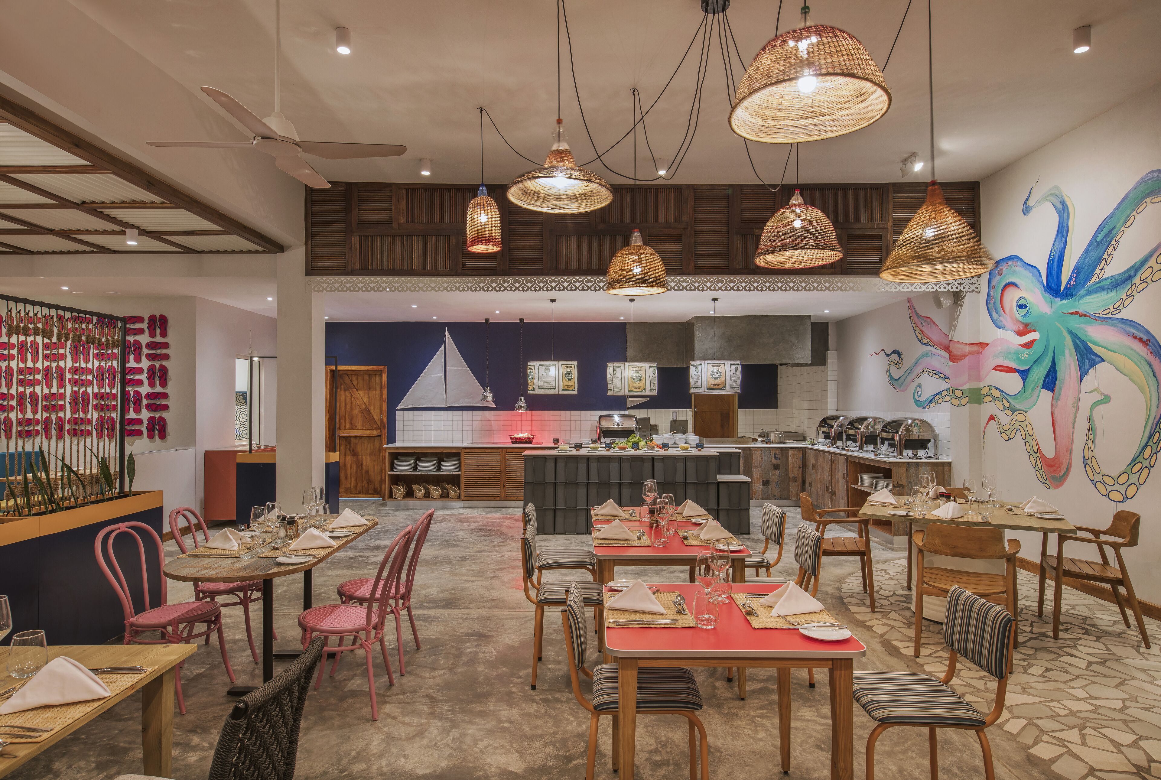 A restaurant with multiple dining tables and chairs, a kitchen in the background, and a colorful mural on the wall.