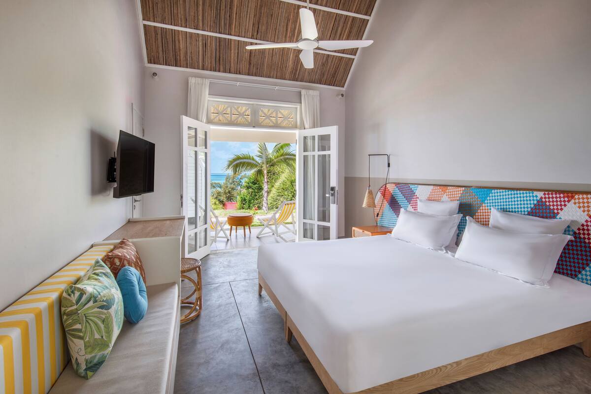 Un dormitorio con cama, televisor, balcón con vista al océano y zona de comedor con sillas.