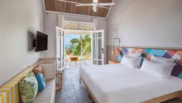 A bedroom with a bed, a TV, a balcony with a view of the ocean, and a dining area with chairs.