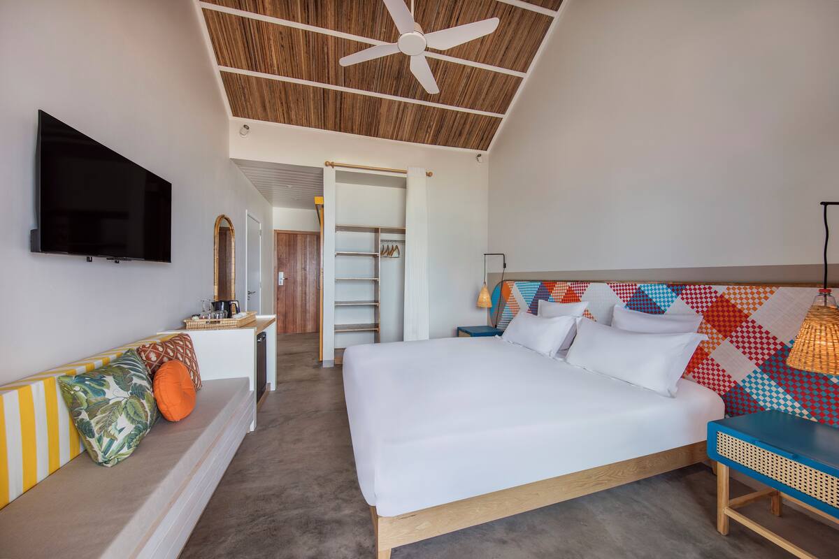 Un dormitorio con una cama, televisión, ventilador de techo y un cabecero colorido.