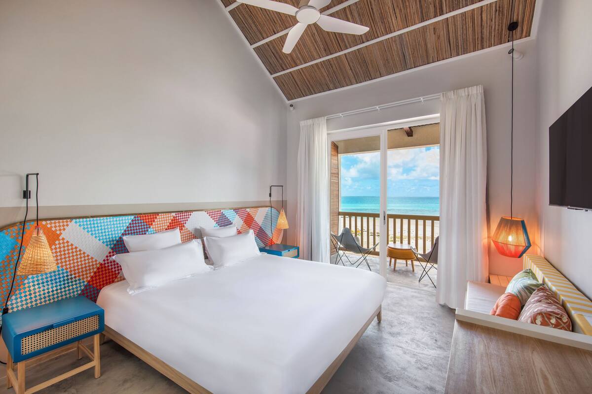 Habitación de hotel con una cama grande, ventilador de techo, balcón con vistas al mar y televisión montada en la pared.