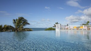 Una piscina infinita azul cristalina con una valla blanca, sillas de descanso, palmeras y vistas al océano.