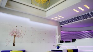 A modern reception area with a white desk, purple and yellow seating, and a decorative wall with a tree and flowers. - VIO Westhoff (Bandung)