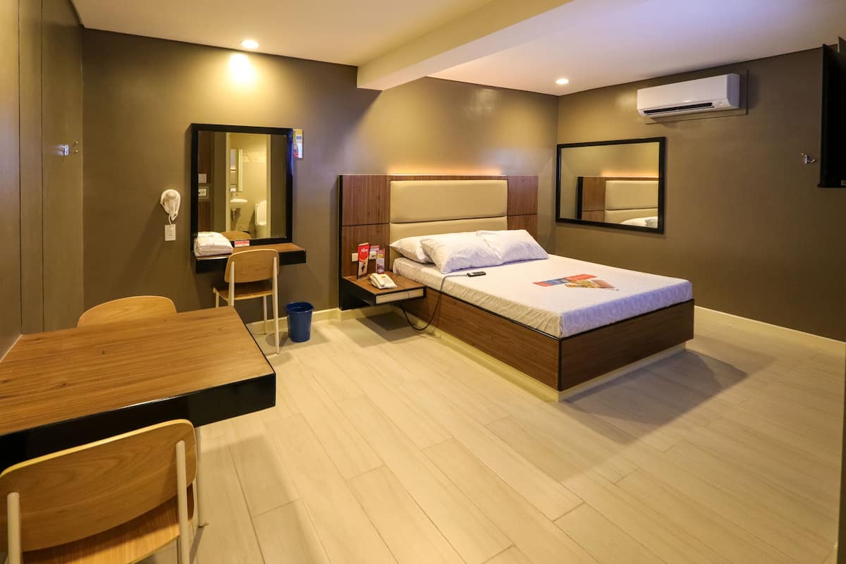 A hotel room with a bed, a desk, a chair, a mirror, and an air conditioning unit.