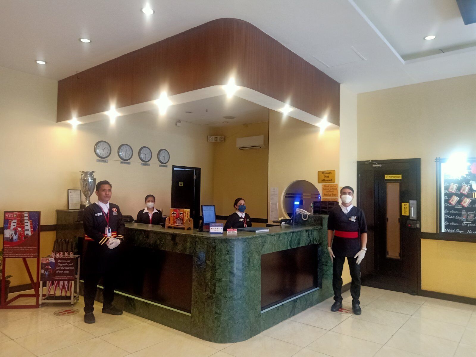 Un mostrador de recepción de hotel con diseño curvo, varios relojes en la pared y un letrero que indica que no se permite el acceso a menores.