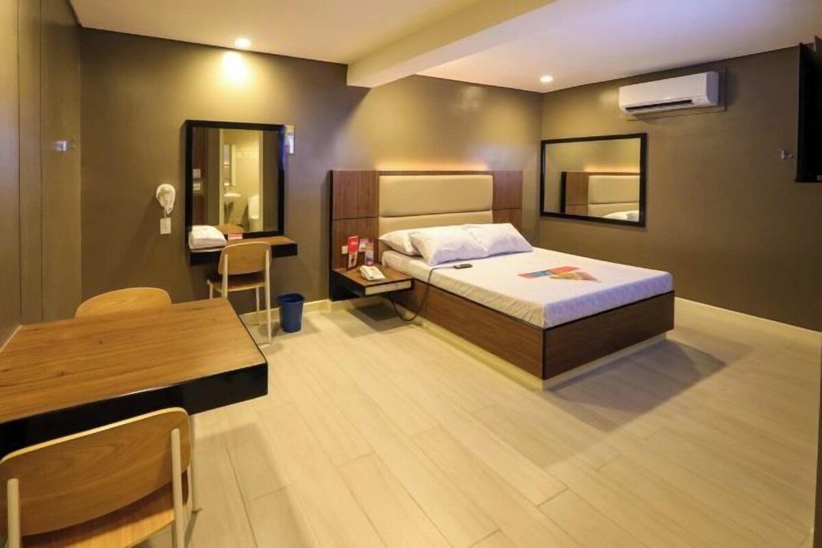 A hotel room with a bed, a desk, a chair, a mirror, and an air conditioning unit.