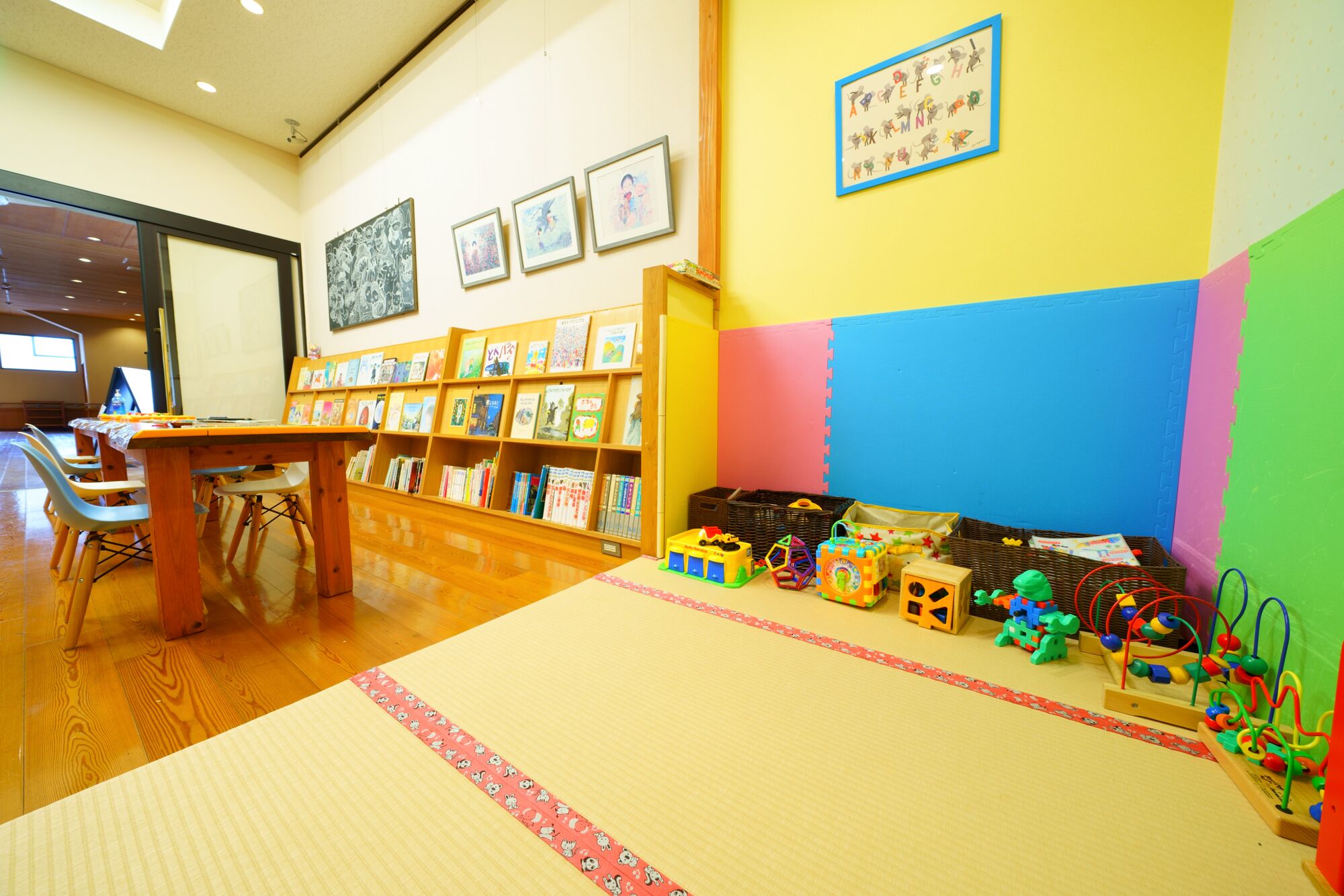 A library with wooden bookshelves, a reading table, and colorful toys on the floor.