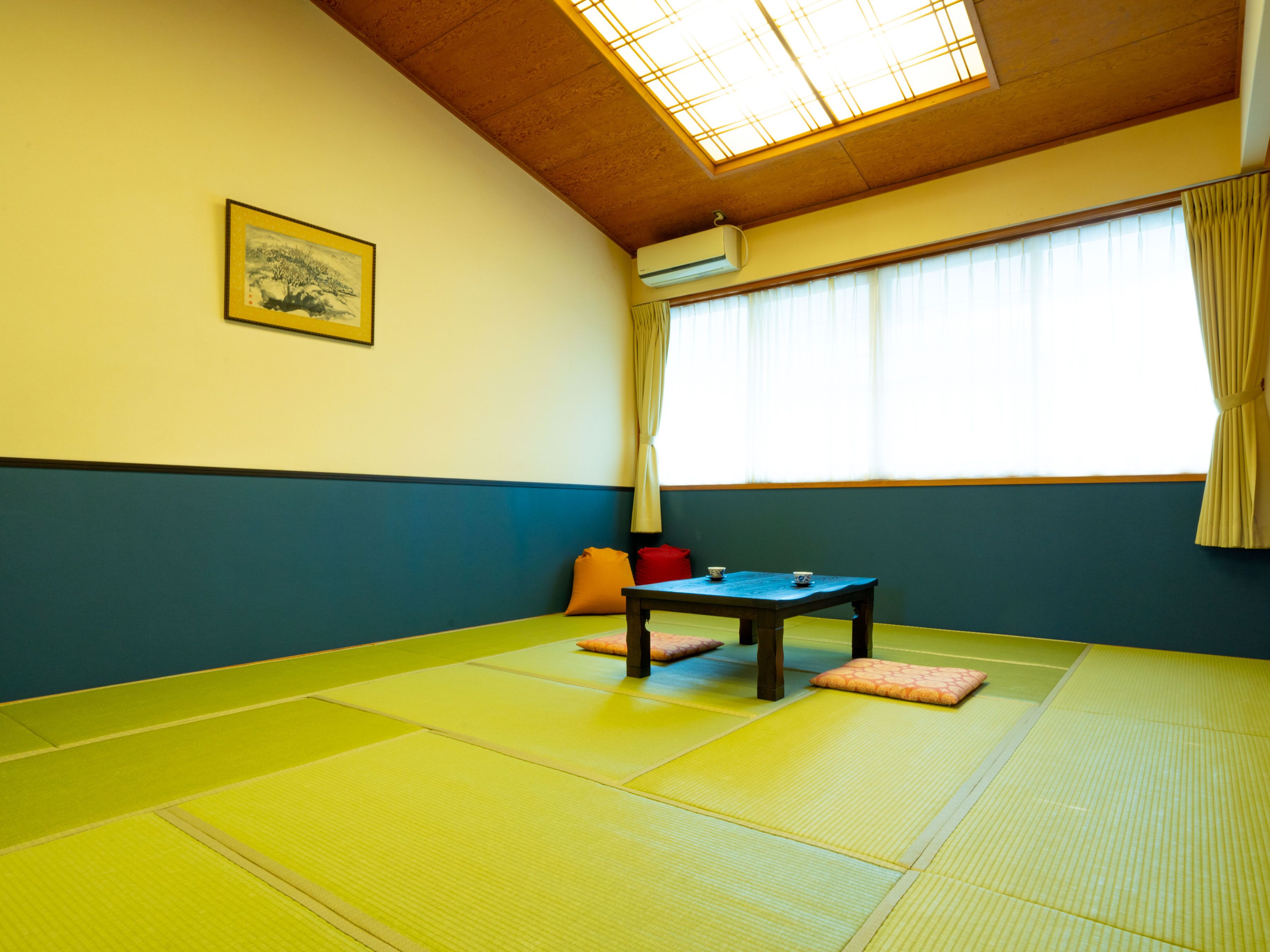 A tatami mat room with a low table, cushions, a framed picture, and a large window with curtains.