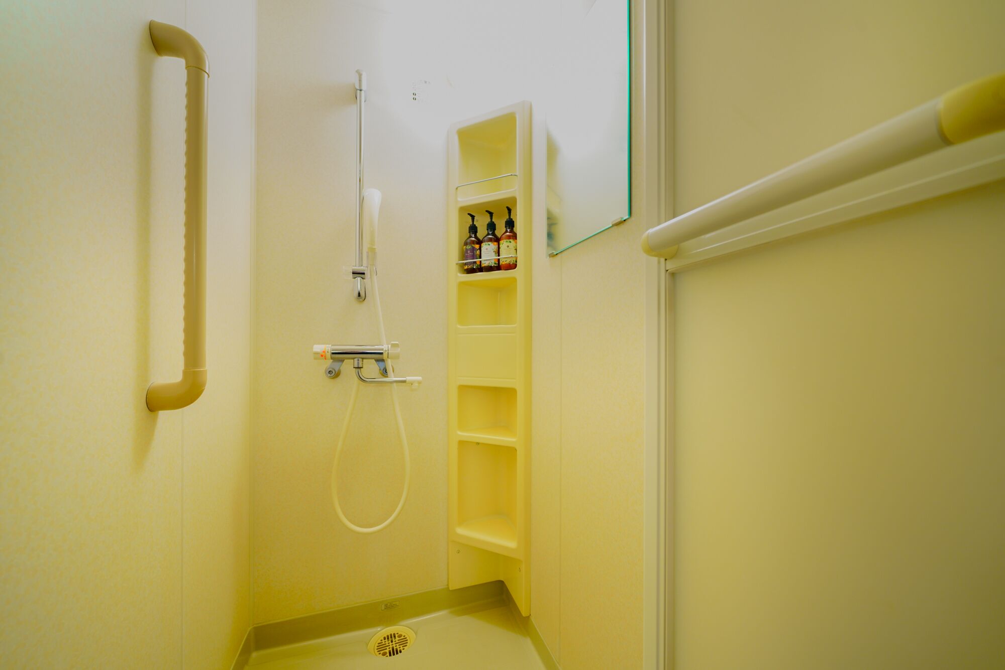 A small, narrow bathroom with a shower, a handrail, and a shelf with toiletries.