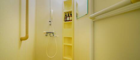 A small, narrow bathroom with a shower, a handrail, and a shelf with toiletries.