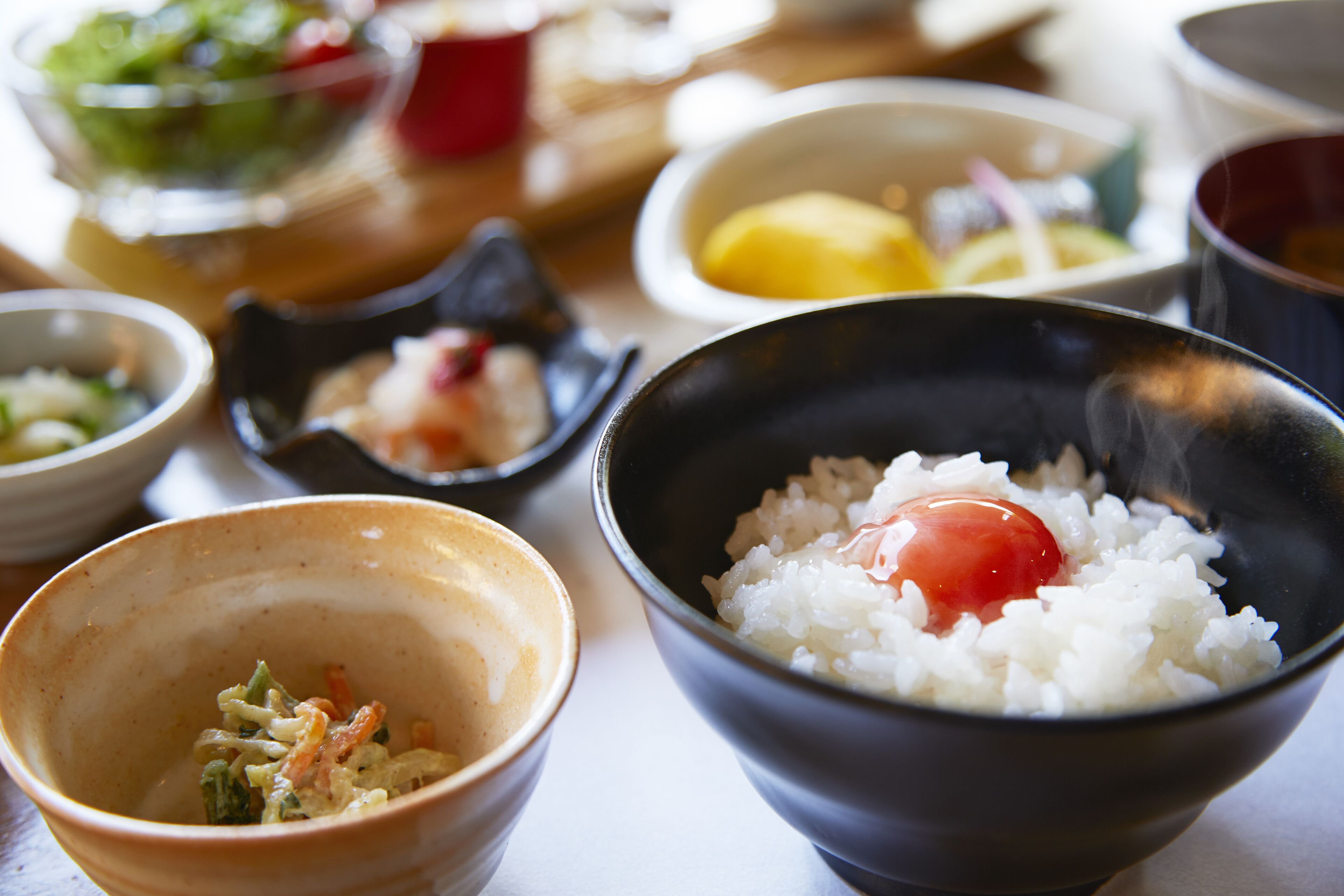 A variety of bowls containing different foods, including rice with a raw egg, vegetables, and possibly some pickled items.