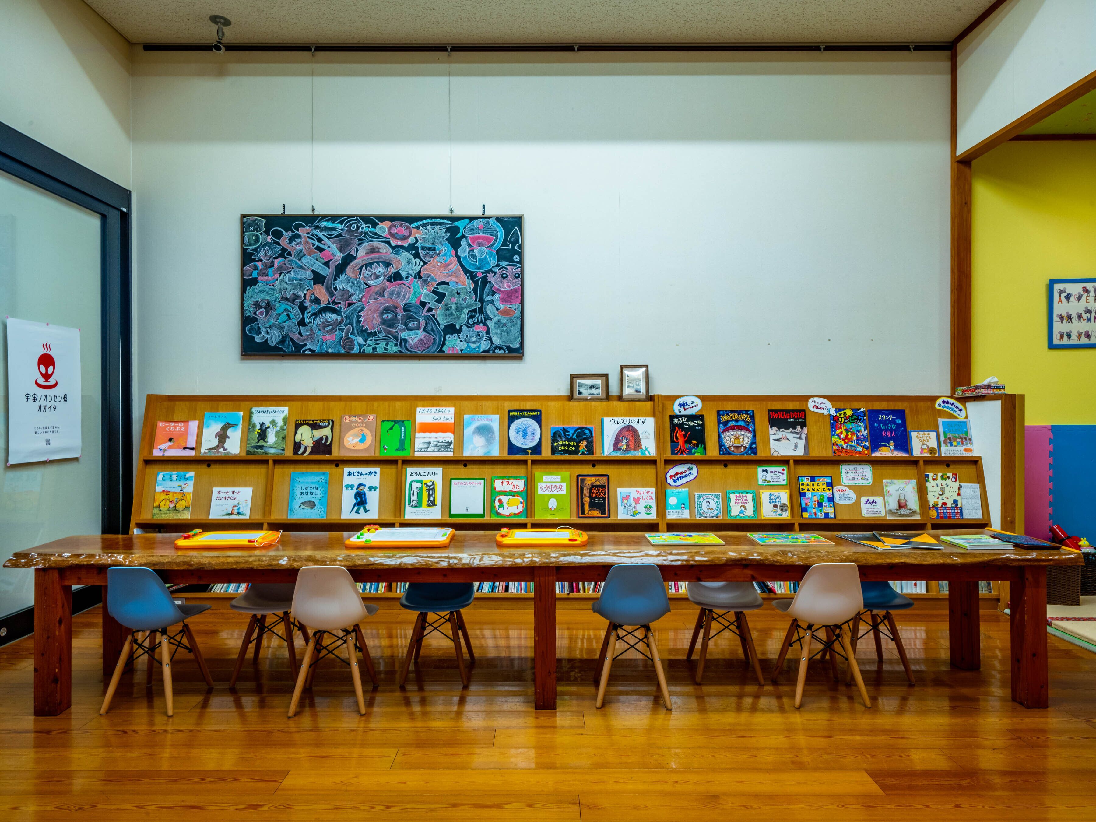Una biblioteca con una larga mesa de madera y varias sillas. Se observan estanterías con libros y una obra de arte colorida.