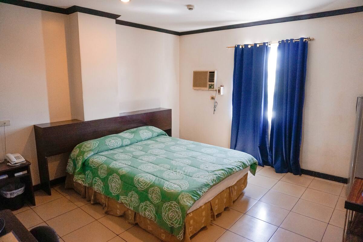 A hotel room with a single bed, a bedside table with a phone, and a wall-mounted air conditioning unit.