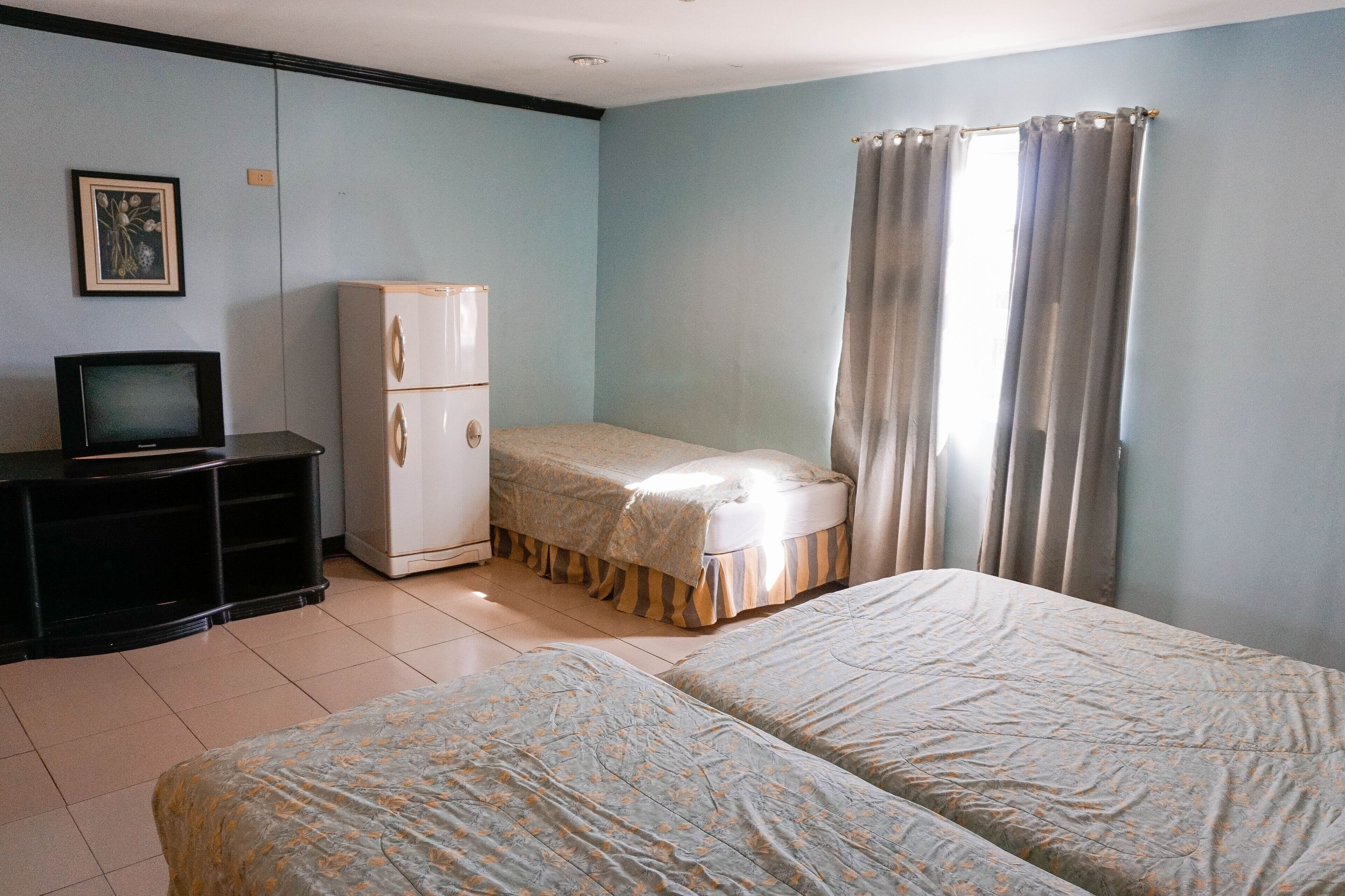 A bedroom with two beds, a television, a refrigerator, and a window with curtains.
