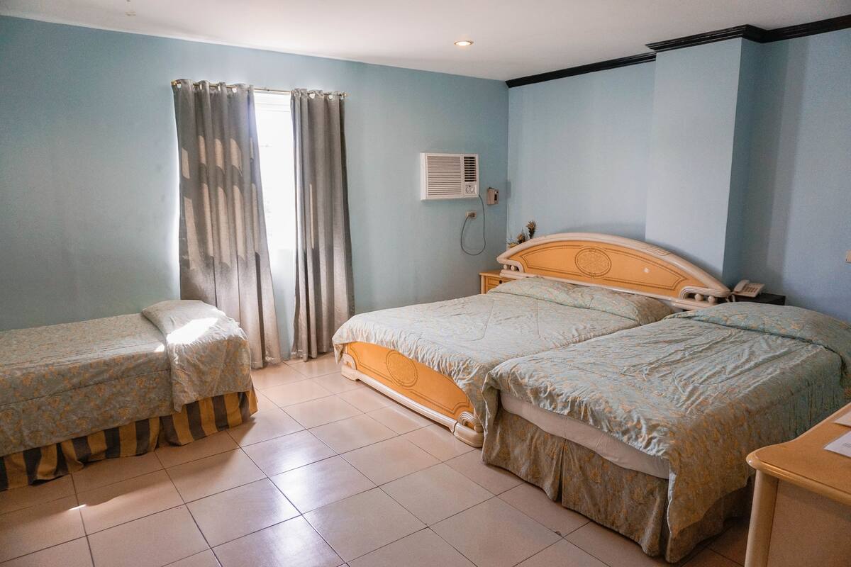 A bedroom with two beds, a window with curtains, an air conditioning unit, and a bedside table.