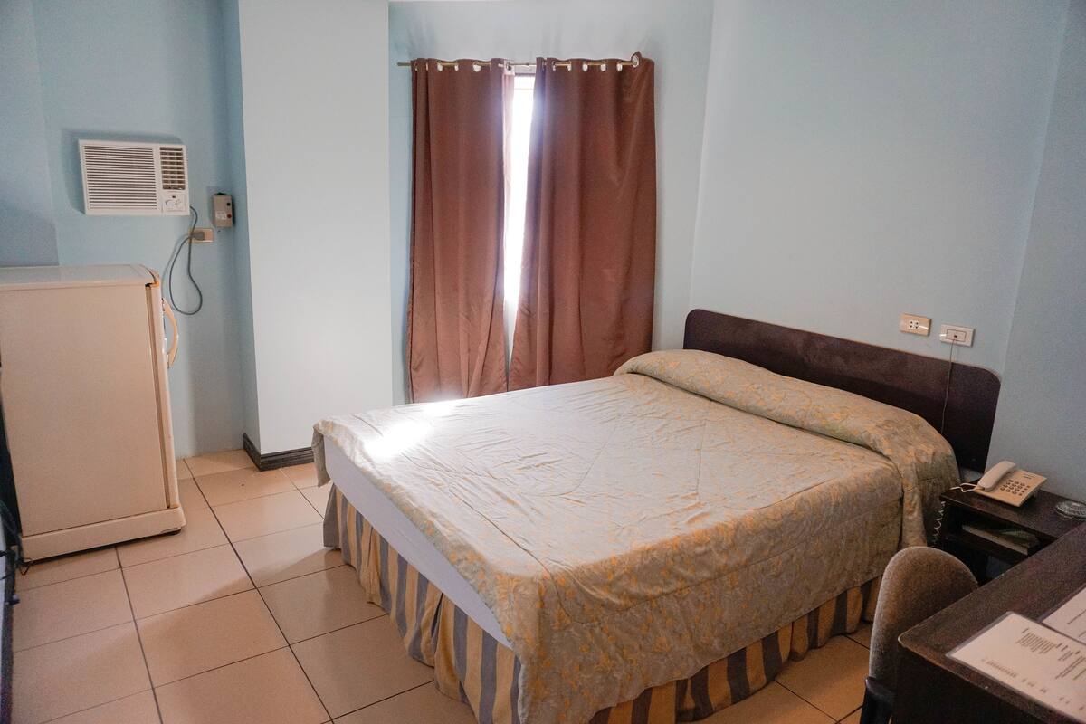 A bedroom with a bed, a desk with a telephone, a wall-mounted air conditioner, and a window with curtains.