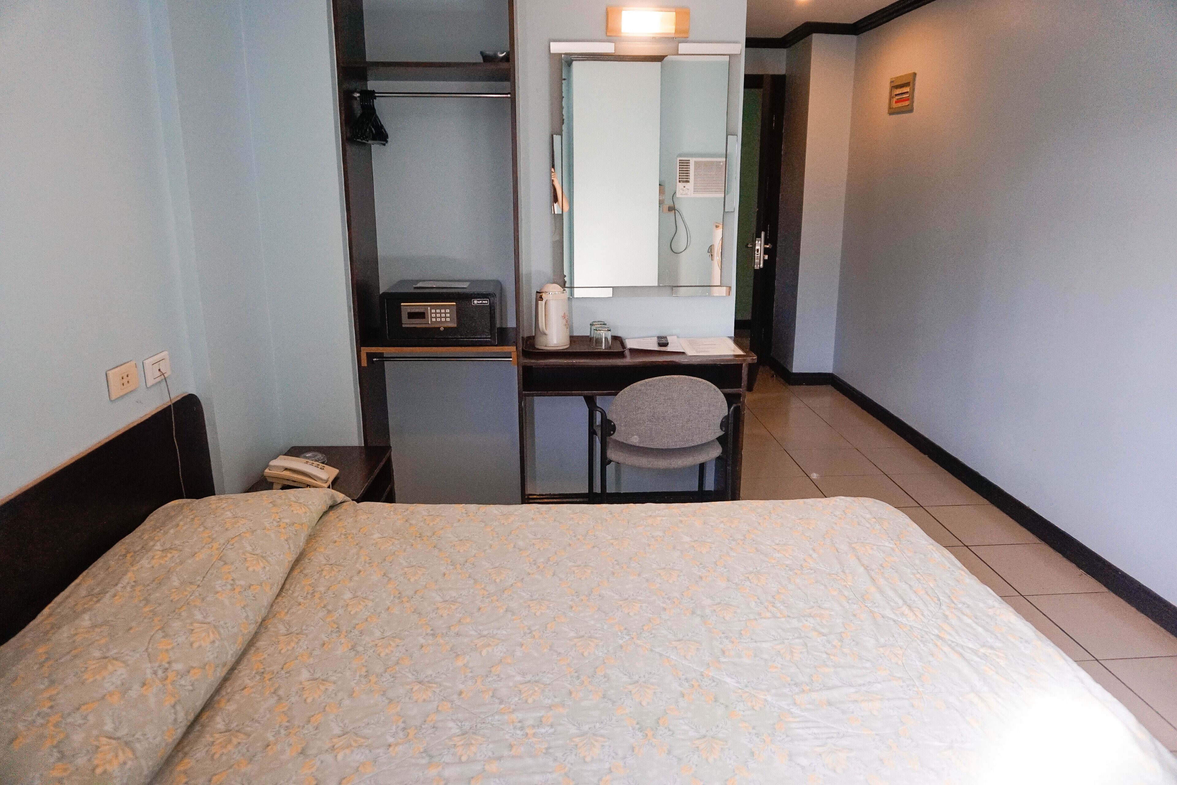 A hotel room with a bed, a desk with a chair, a mirror, a telephone, and a small table with a cup and a book.