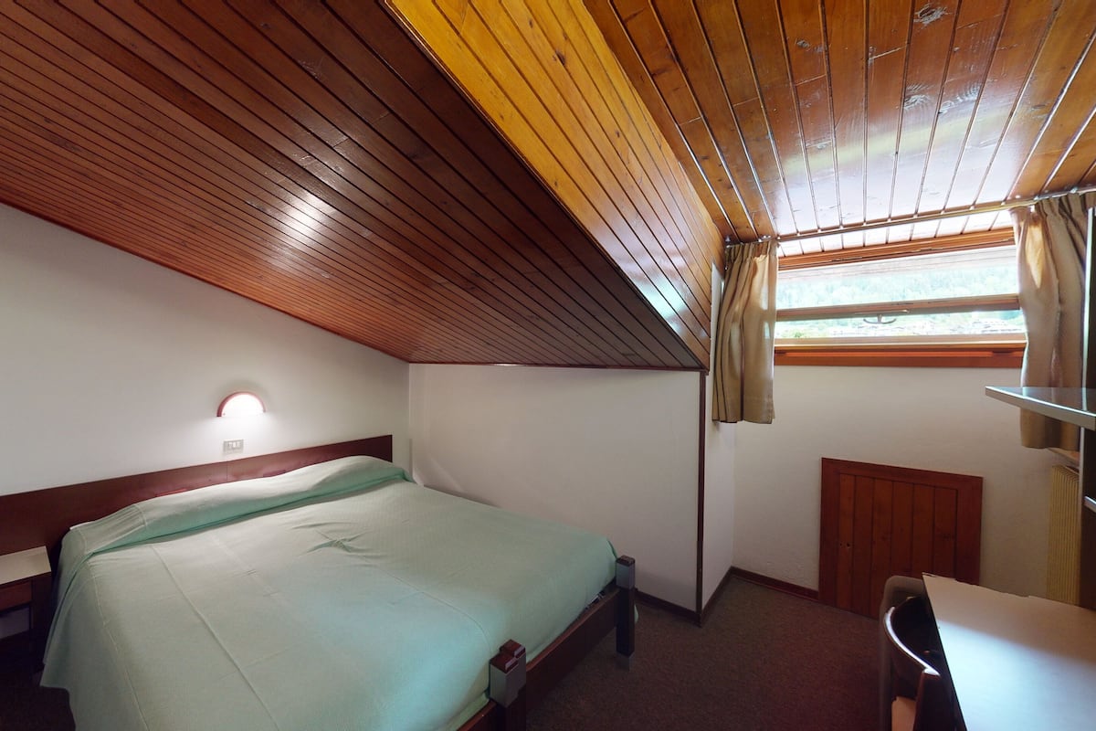 A small, single-bed room with a sloped ceiling, a bedside table, a lamp, and a window with curtains.