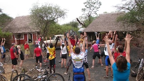 A group of people outdoors, some with bicycles, gathered around a vehicle in a rural setting with thatched-roof structures. - Maasai Simba Camp (Amboseli)