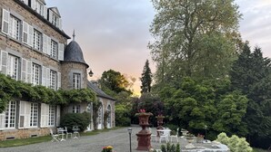 Een historisch stenen gebouw met veel ramen en een opvallende koepel, omgeven door een verzorgde tuin met diverse planten en zitplaatsen.