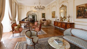 Een statige kamer met een piano, versierde stoelen, een bank, een kroonluchter en een grote spiegel.