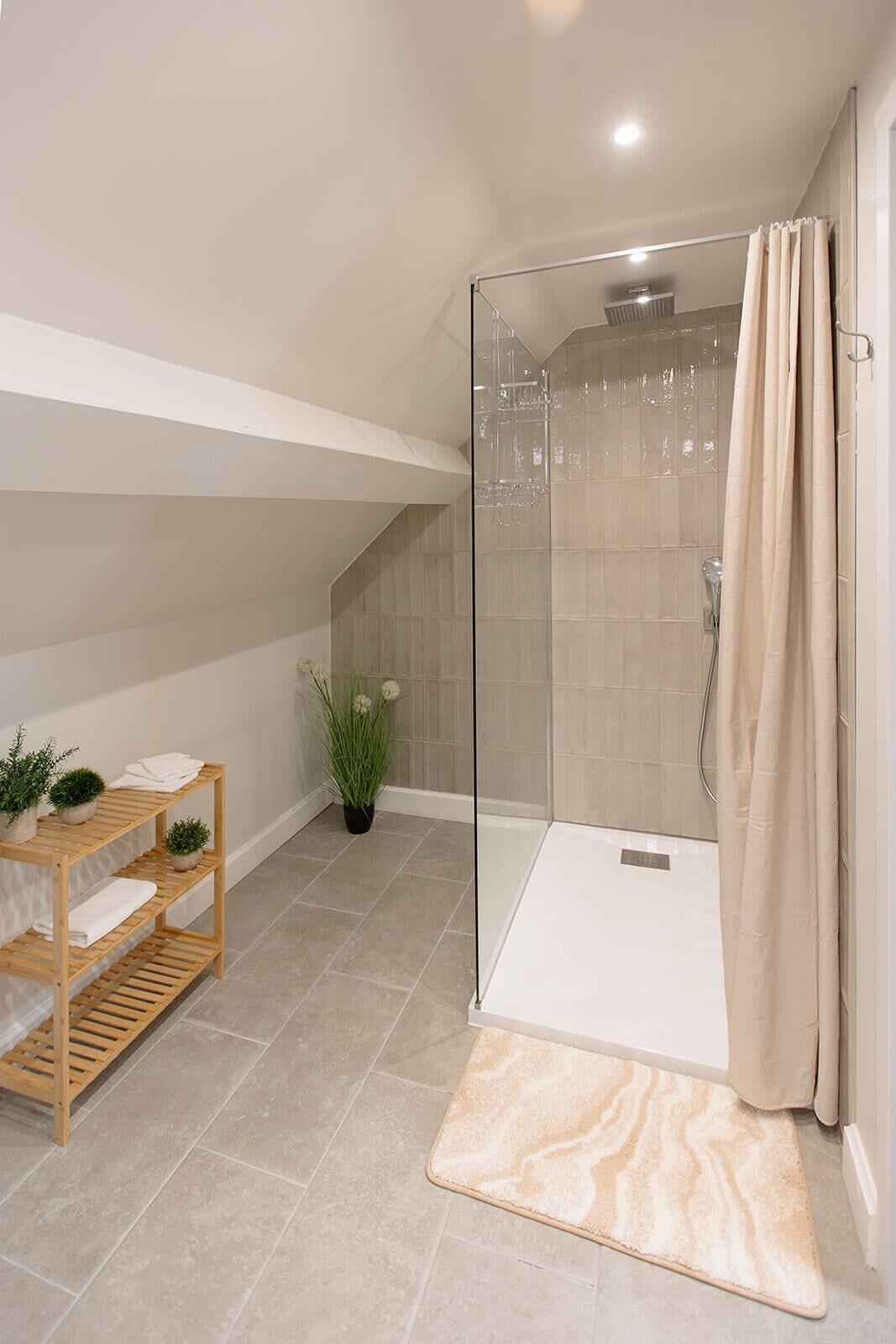 A shower with a glass door, a wooden shelf with towels, a potted plant, and a marble-patterned mat.