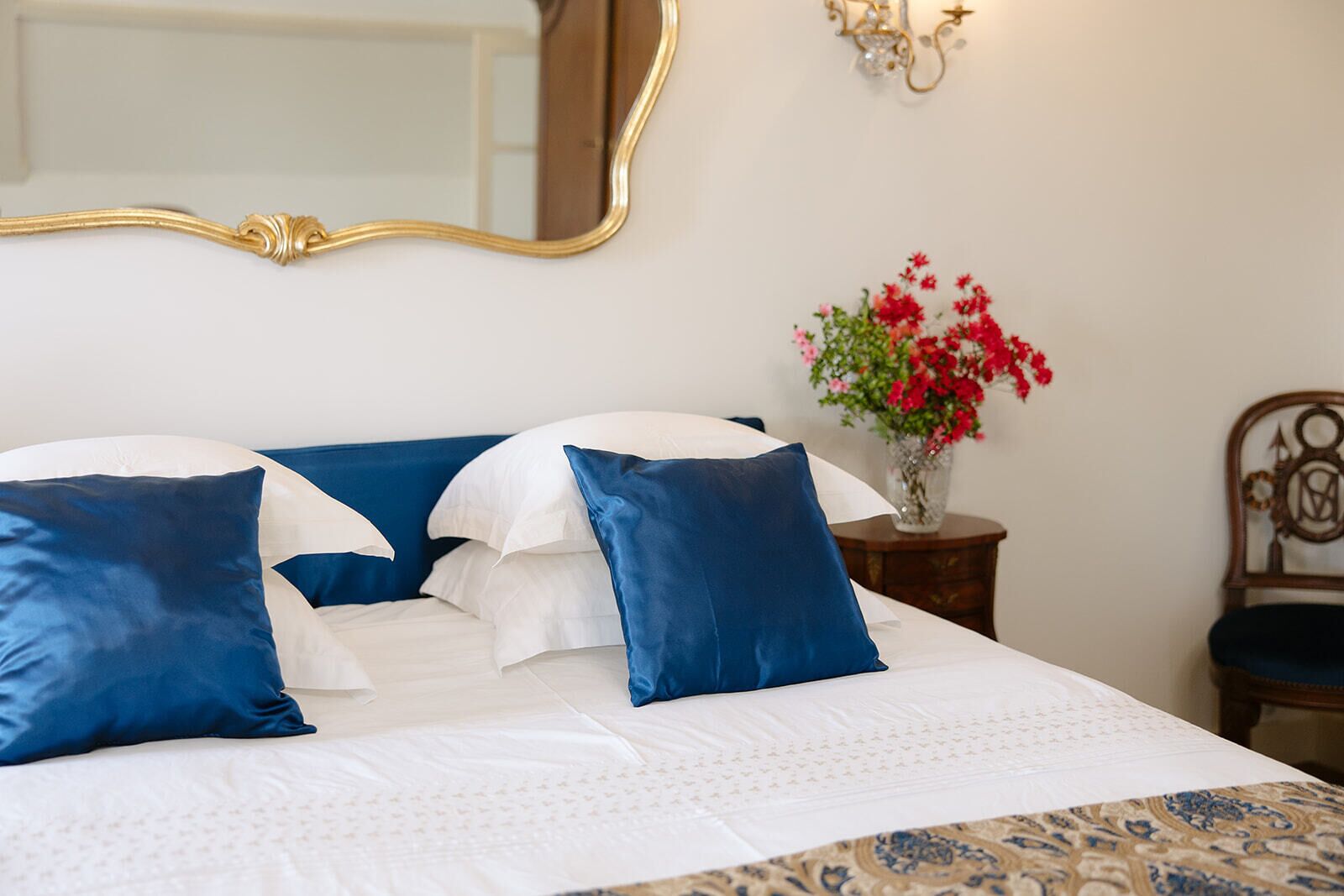 A neatly made bed with white and blue bedding, a mirror above it, and a vase with red flowers on a bedside table.