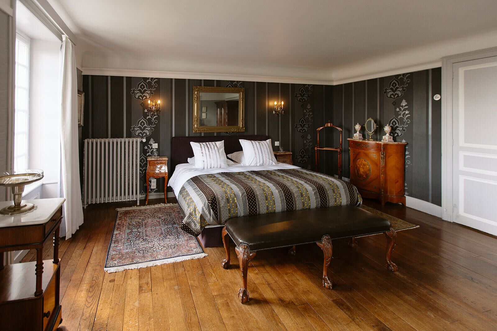 Un dormitorio con una cama grande, un banco de madera, un aparador, un espejo y un radiador.