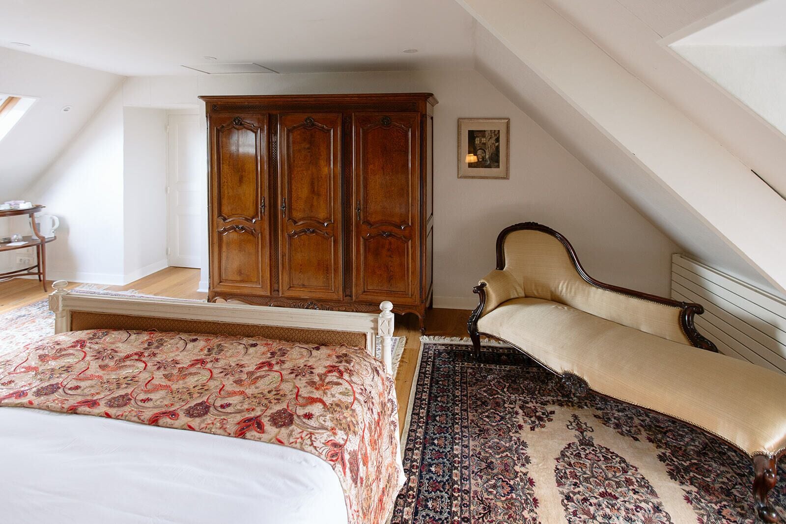 A bedroom with a bed, a sofa, a wooden wardrobe, a patterned rug, and a framed picture on the wall.