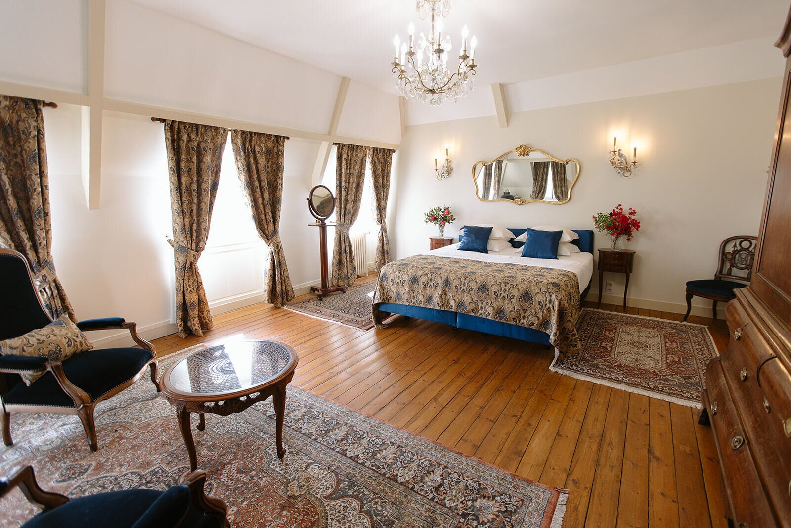 A bedroom with a bed, a round table, a chair, a dresser, a mirror, and a chandelier.
