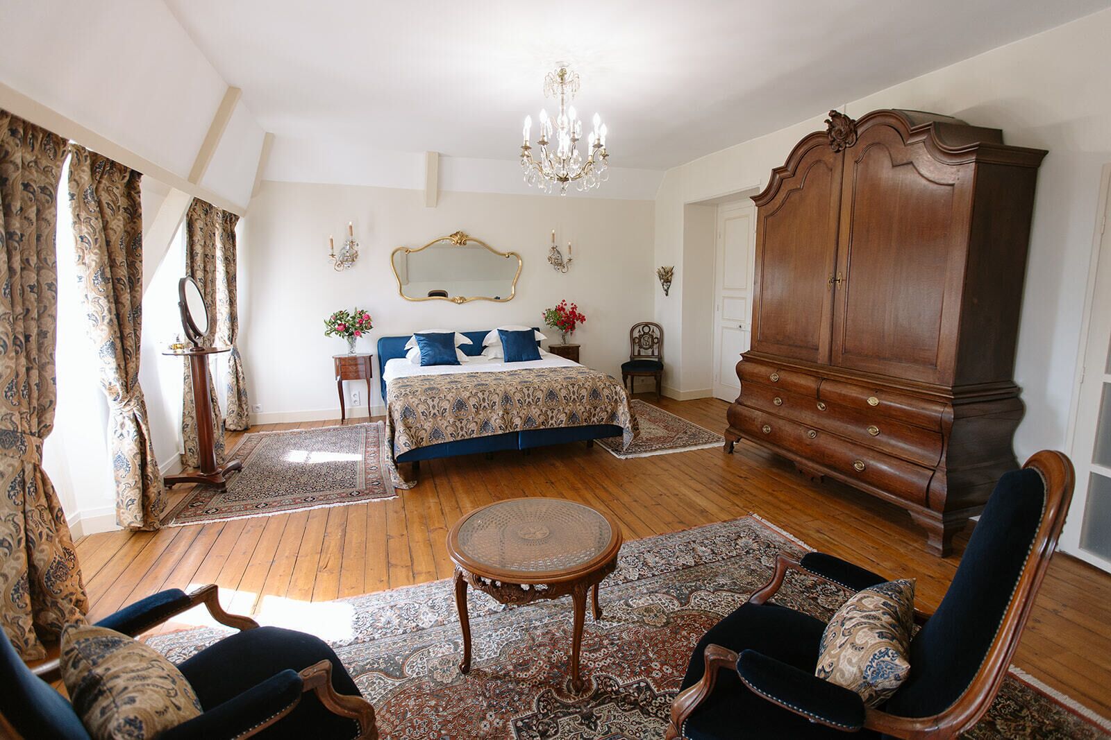 A bedroom with a large bed, a wooden wardrobe, a chandelier, and a sitting area with a table and chairs.