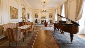 Een statige kamer met een vleugelpiano, versierde stoelen, een kroonluchter en een open haard.