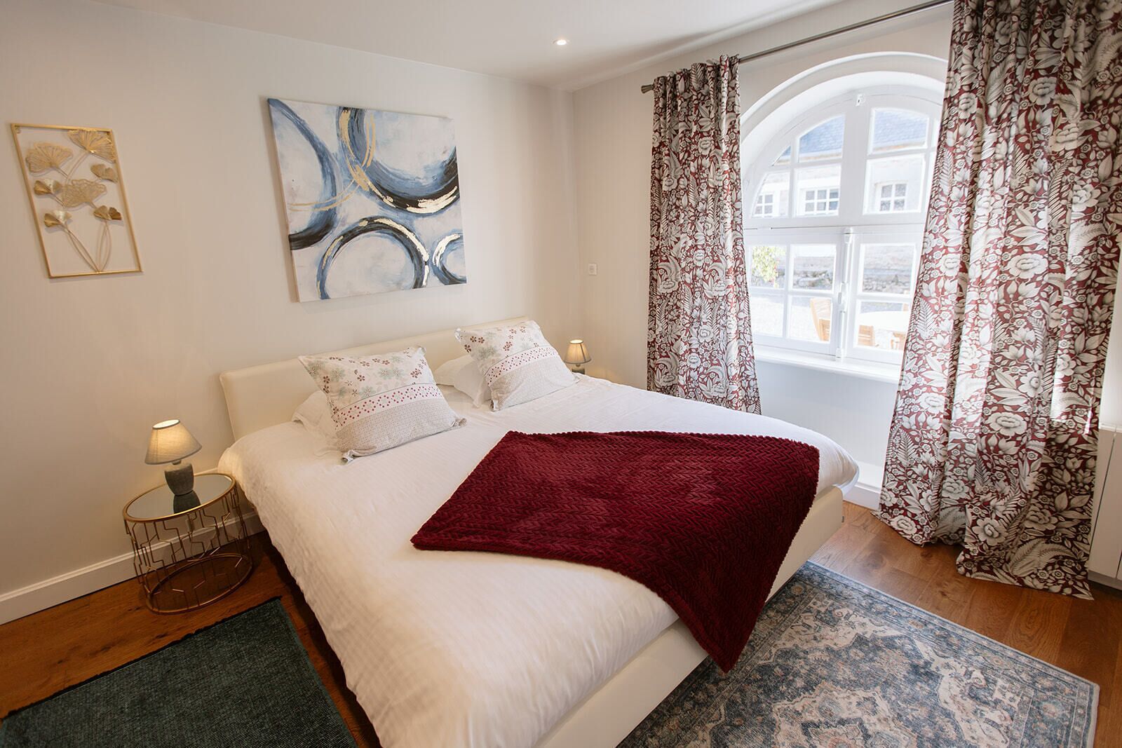 A bedroom with a large bed, two bedside lamps, a red throw blanket, a blue abstract painting, and floral curtains.