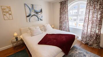 A bedroom with a large bed, two bedside lamps, a red throw blanket, a blue abstract painting, and floral curtains.