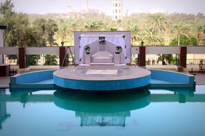 A poolside area with a stage set up for an event, surrounded by palm trees and a view of a tower in the background. - Aifu Resort (Alexandria)